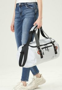 Sac de sport blanc avec des poignées noires, des accents orange et des zippers. Dispose de doubles poignées et d'un compartiment zippé sur un côté.