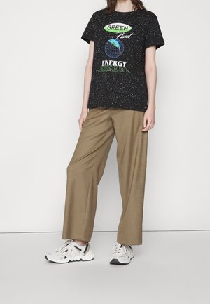 Sort t-shirt med et spættet mønster og grafisk tekst i grøn, blå og hvid. Bred beige bukser og hvide sneakers fuldender looket.