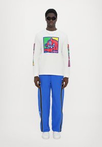 Hvit langermet skjorte med fargerik grafisk design, kombinert med blå joggebukser med sidestriper, og hvite sneakers. Sorte solbriller brukes.