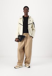 Veste zippée Nike en crème clair avec des accents noirs, t-shirt noir, pantalon large beige et baskets sportives beiges, tenant une pochette noire.