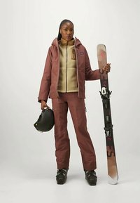 Snowboarder, der eine burgunderfarbene wasserdichte Jacke und passende Hose trägt, hält einen schwarzen Helm und ein Snowboard mit schwarzen und roten Akzenten.