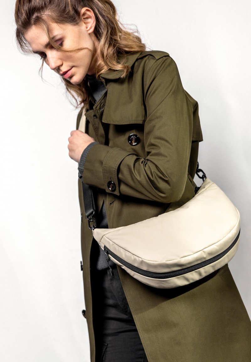 Beige, gepolsterte Umhängetasche mit einer geschwungenen Form und schwarzen Reißverschlüssen, getragen über einem olivgrünen Trenchcoat.