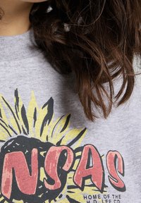 Ingrandimento di un felpa grigia con una grafica di girasole e grandi lettere rosse "NSAS" sul petto, parzialmente coperta da capelli castani mossi.