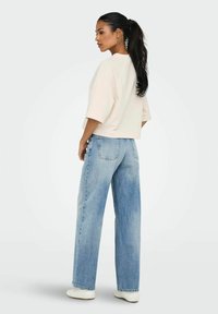 Frau mit einem langen, dunklen Pferdeschwanz, die ein weißes Oberteil, helle, weit geschnittene Jeans, weiße Schuhe und Hoops-Ohrringe trägt und in einem neutralen Raum steht.
