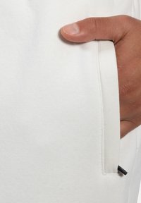 Biała materiałowa kieszeń o gładkiej, eleganckiej fakturze, z czarnym akcentem po boku. Ręka umieszczona wewnątrz kieszeni.