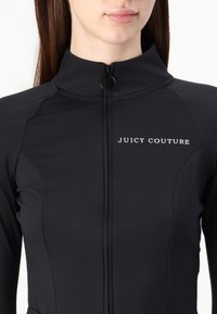 Čierna zipová bunda vyrobená z hladkej tkaniny; má vysoký golier a biele logo "JUICY COUTURE" na hrudi. Zipsový detail na krku.