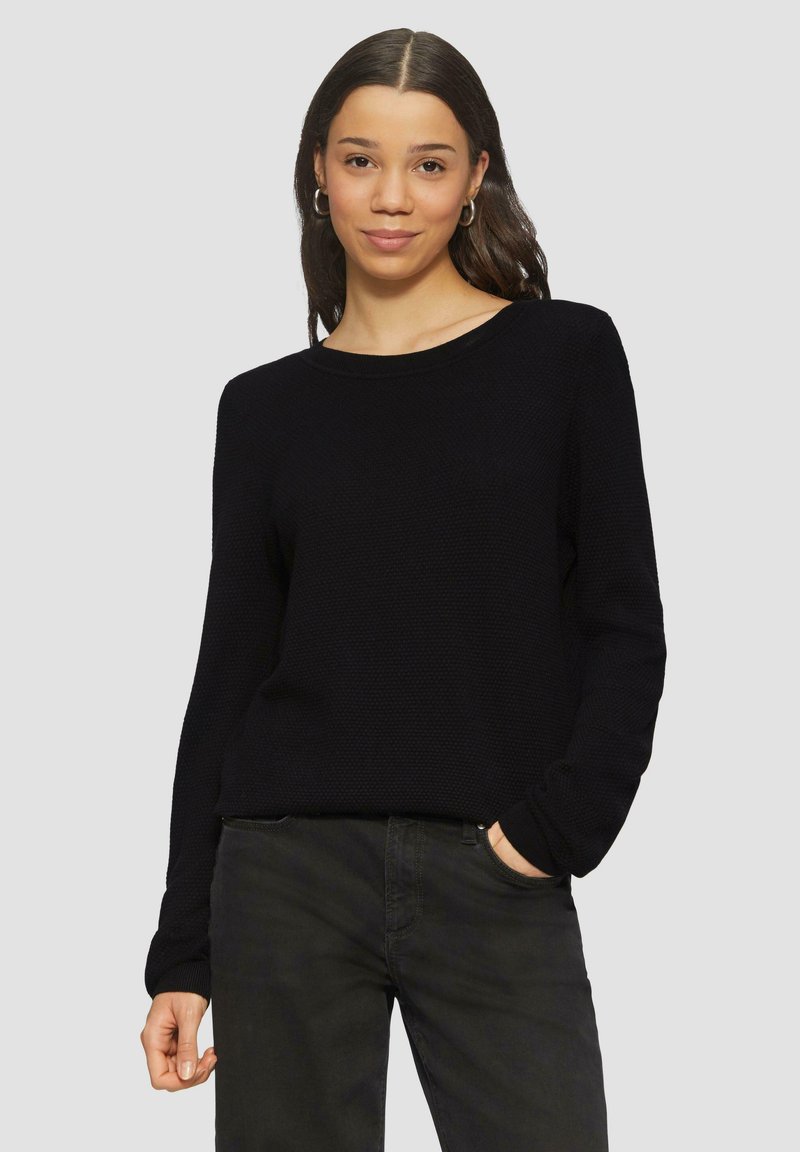 Pull en maille noir avec un col rond et des manches longues, arborant un motif texturé. Associé à un jean foncé, les mains placées de manière décontractée dans les poches.