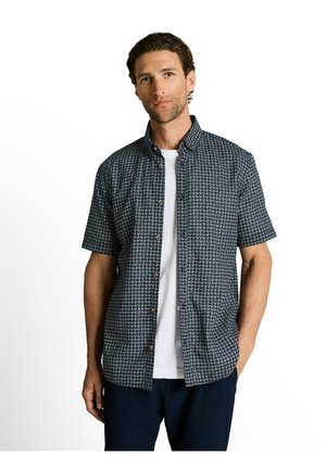 Mann trägt ein kurzärmeliges, marineblau gemustertes Hemd über einem weißen T-Shirt und steht mit einer Hand in der Tasche.