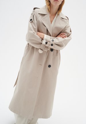 Woman wearing a beige double-breasted trench coat with black buttons, standing with arms crossed against a white background.