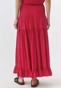 Roter Maxirock mit gerafftem Taillenbund, gestuften Rüschen am Saum und fließendem Stoff. Getragen mit flachen schwarzen Sandalen.