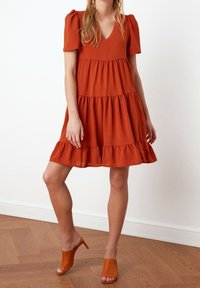 Rostfarbendes Baumwollkleid mit Puffärmeln, gestuften Design, Rüschensaum und V-Ausschnitt. Trägt dazu passende Sandalen mit offener Zehe.