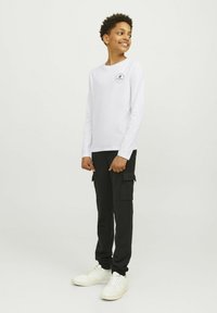 Vit långärmad skjorta med en liten svart logotyp, tillsammans med svarta cargobyxor med sidofickor, och vita sneakers.