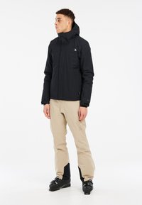 Giacca nera impermeabile con cappuccio, tasche laterali e logo sul petto abbinata a pantaloni beige e stivali da sci neri.