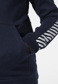 Sudadera azul marino con un bolsillo tipo canguro; presenta un detalle de rayas plateadas en la manga y puños acanalados para añadir textura.
