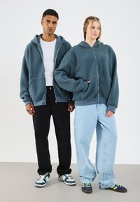 Twee personen dragen vergelijkbare oversized, rits-hoodies van zacht, gestructureerd, donkerblauw-groen stof, gecombineerd met casual broeken en sneakers.