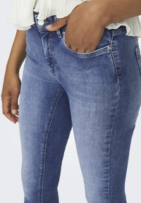 Personne portant un jean skinny bleu délavé avec une main dans la poche et un haut blanc à volants partiellement visible.