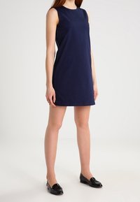 Námořnické šaty bez rukávů s kulatým výstřihem, délka ke kolenům a hladká tkanina. Modelka má na sobě černé baleríny.