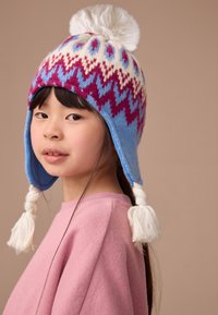 Cappello a maglia con base blu, decorato con motivi a zigzag crema e marrone, sormontato da un pompon bianco e caratterizzato da nappe laterali.