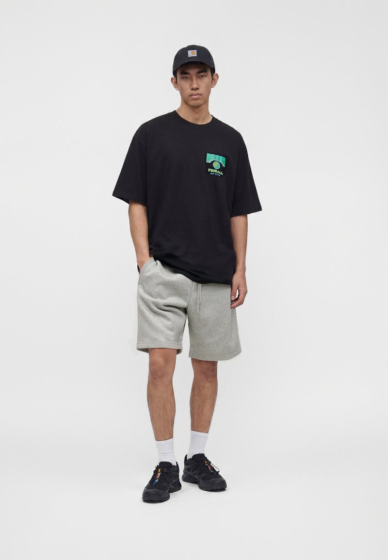 Junger Mann trägt schwarzes T-Shirt mit Grafik, graue Shorts, schwarze Sneakers mit Socken und eine dunkelblaue Kappe vor schlichtem grauem Hintergrund.