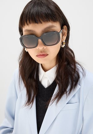 Donna con capelli lunghi e scuri e frangia indossa grandi occhiali da sole neri, orecchini a grappolo di perle, camicia bianca con colletto, maglione nero e blazer azzurro chiaro.