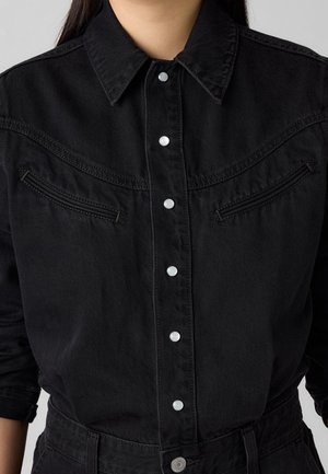 Frau trägt ein schwarzes Jeanshemd mit silbernen Druckknöpfen und Reißverschluss-Brusttaschen, hochgekrempelte Ärmel und passende schwarze Hose.