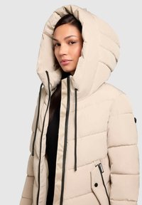 Vrouw in een beige pufferjack met een oversized capuchon en zwarte rits- en trekkoorddetails tegen een lichtgrijze achtergrond.