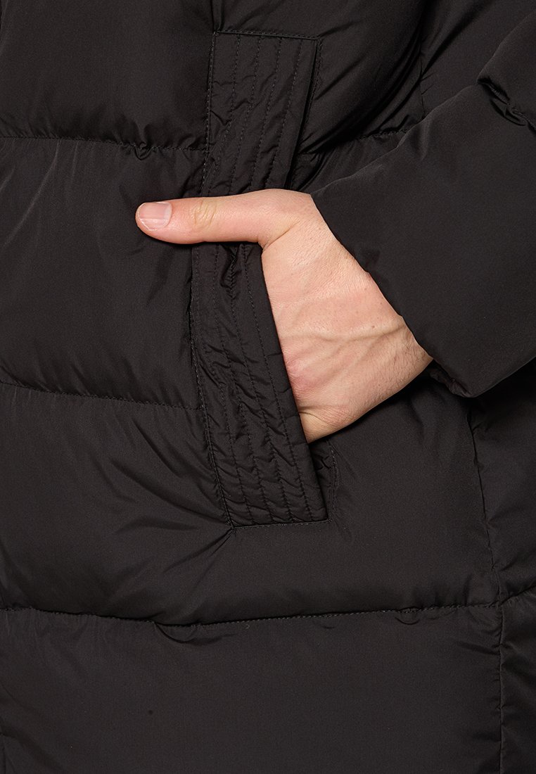 Zwarte pufferjacksectie met gestikte details en een zijzak, handmatig ingeshoven. Gladde textuur en zichtbare isolatie.