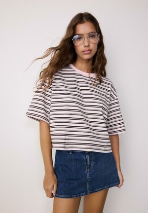 Giovane donna con capelli lunghi e mossi che indossa grandi occhiali da aviatore trasparenti, una maglietta rosa a righe larga e una minigonna in denim blu.