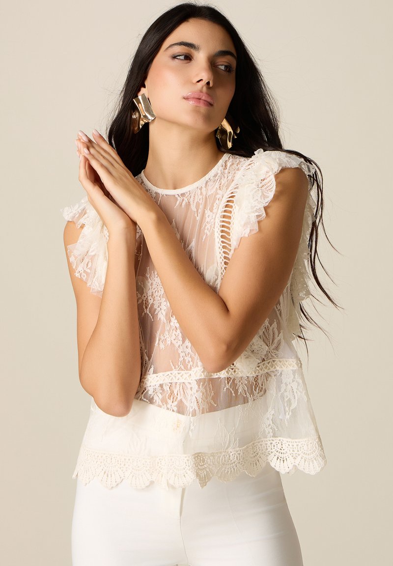 Mujer con cabello largo y oscuro, lleva grandes pendientes dorados y un top sin mangas de encaje blanco, con las manos entrelazadas cerca del rostro.