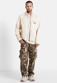 Lichtbeige gekraagde jas met rits en borstzak; gecombineerd met bruine camouflage cargo broek met meerdere zakken. Witte en beige sneakers.