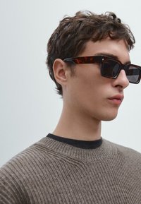 Occhiali da sole tartarugati con forma rettangolare, lenti scure e una montatura spessa, abbinati a un maglione a coste marrone e un colletto nero.