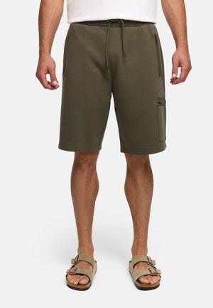 Uomo con pantaloncini verde oliva con coulisse e tasche con zip e sandali beige, in piedi davanti a uno sfondo semplice.