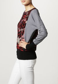 Sweat-shirt gris à manches longues avec un motif floral rouge sur le devant, une ceinture noire et une coupe décontractée. Détails avec boutons sur la manche.
