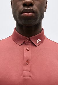 Άτομο με σκουρόχρωμο δέρμα που φοράει πουκάμισο πόλο σε αποχρώσεις ρόδου-σκονισμένο ροζ με κουμπιά και μικρό λευκό λογότυπο στο γιακά, σε φωτεινό φόντο.