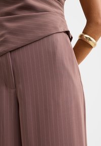 Vrouw draagt een mauve pinstripe rok en top, met een gouden hamerslag armband aan haar pols tegen een witte achtergrond.