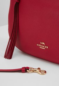 Sac à main en cuir rouge avec une finition texturée, des accessoires dorés et plusieurs longs pompons. Comprend une bandoulière amovible avec un fermoir doré.