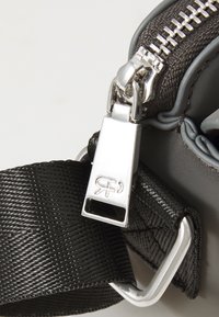 Γκρι τσάντα αξεσουάρ με ασημί φερμουάρ, μαύρη υφασμάτινη λωρίδα και μεταλλική λεπτομέρεια με λογότυπο στον κρίκο του φερμουάρ.