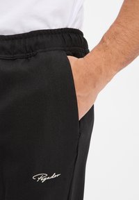 Schwarze Hosen im Fischgrätenmuster mit elastischem Bund und Seitentaschen. Verfügt über ein dezentes Logo in weißer Stickerei.