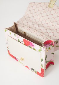 Sac à main à motif floral avec un extérieur en toile rose pâle, rabat crème, accessoires dorés et une poche intérieure zippée.
