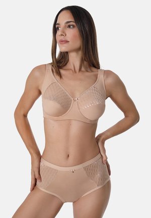 Frau trägt beigen BH und passende hoch taillierte Slips mit strukturierten Netzpaneelen, steht mit den Händen in der Hüfte und schaut nach links.