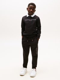 Μαύρο φούτερ με λευκό γιακά και λογότυπο "TOMMY HILFIGER", συνδυασμένο με μαύρους αθλητικούς παντελόνες που έχουν εκτύπωση λογότυπου στο πλάι, φορεμένο με λευκά αθλητικά παπούτσια.