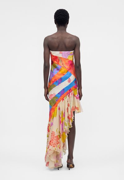 Γυναίκα, ειδωμένη από πίσω, φοράει ασύμμετρο πολύχρωμο φόρεμα χωρίς τιράντες με λουλουδένιο και ριγωτό σχέδιο και βολάν στο τελείωμα, στεκόμενη μπροστά σε απλό φόντο.