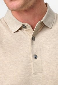 Beige pikétröja i textil med struktur, tre mörka knappar på placket och en klassisk kragsdesign.