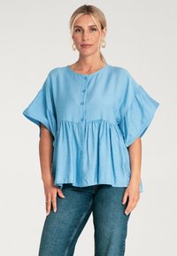Jasnoniebieska bluzka z krótkim rękawem o okrągłym dekolcie, z guzikami z przodu i marszczonym dołem, wykonana z gładkiego materiału; zestawiona z ciemnymi dżinsami.
