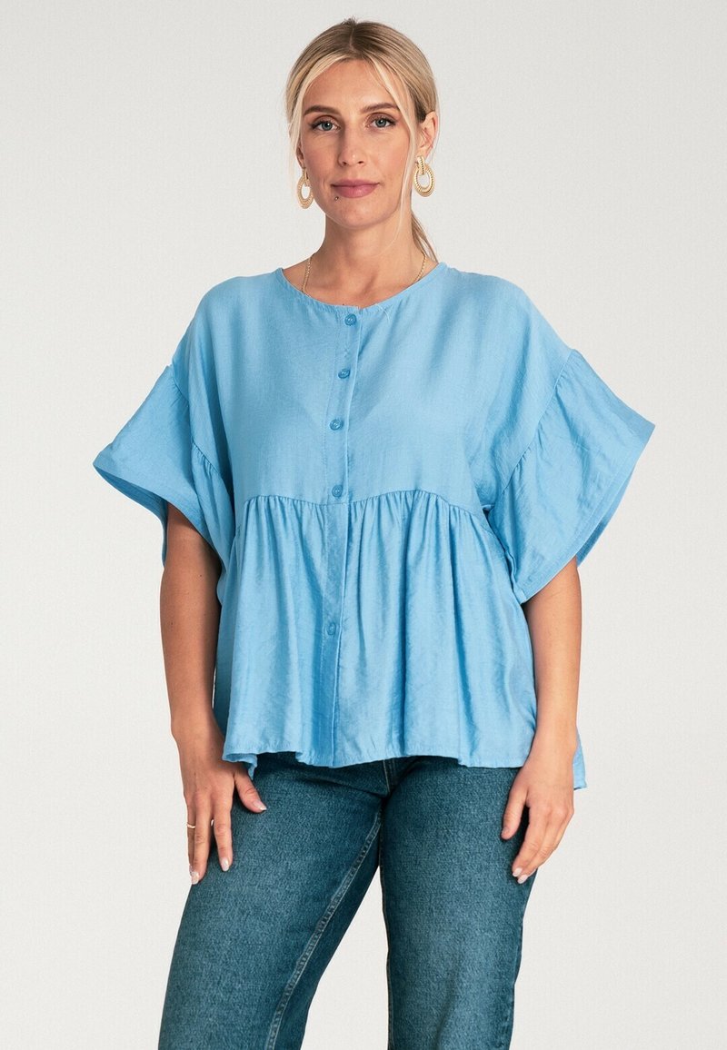 Jasnoniebieska bluzka z krótkim rękawem o okrągłym dekolcie, z guzikami z przodu i marszczonym dołem, wykonana z gładkiego materiału; zestawiona z ciemnymi dżinsami.