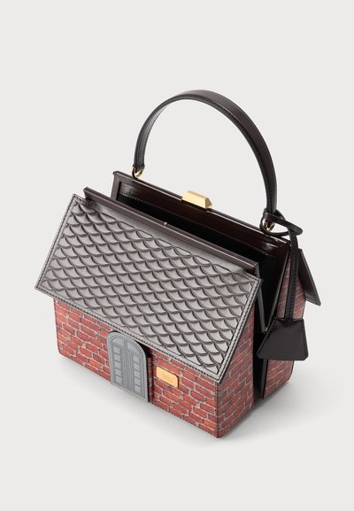 Sac à main en forme de maison en briques avec un rabat de toit en tuiles grises, une porte grise et une poignée noire, présenté ouvert sur fond blanc.