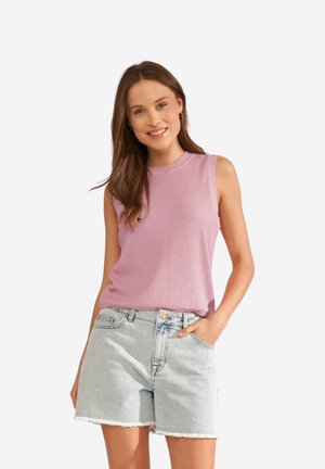 Femme souriante aux cheveux bruns portant un haut sans manches côtelé lavande et un short en jean clair avec des ourlets effilochés, posant avec une main dans la poche.