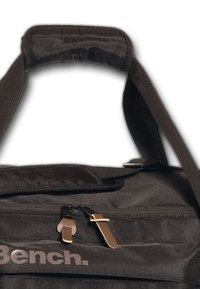 Sac de sport en tissu noir avec fermeture éclair double, poignée supérieure rembourrée et bandoulière ajustable. Présente un logo gris sur le côté.