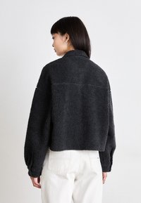 Jachetă neagră, pufoasă, de tip cropped, cu un finisaj texturat, având un guler și mâneci lungi, ușor umflate. Purtați cu pantaloni de culoare deschisă.