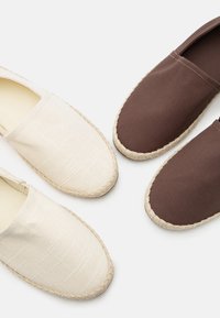 Δύο ζευγάρια εσπαντρίγιες slip-on, το ένα σε μπεζ λινό και το άλλο σε καφέ ύφασμα, το καθένα με πλεκτές σόλες από γιούτα, σε λευκό φόντο.